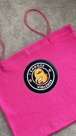 یک تاپ یا کراپ زنانه صورتی با بندهای نازک اسپارگتی. در مرکز سینه تاپ، یک پچ دایره‌ای سیاه با حاشیه سفید و طرح یک جوجه کارتونی زرد رنگ وجود دارد که قلبی قرمز در دست دارد و زیر آن به انگلیسی نوشته شده: "I CHOOSE VIOLENCE" (من خشونت را انتخاب می‌کنم). در اطراف طرح جوجه، جمجمه‌های کوچکی نیز دیده می‌شود. پارچه تاپ به نظر بافت‌دار و راه راه عمودی است و از نمای نزدیک عکاسی شده است .