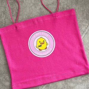 یک تاپ یا کراپ زنانه صورتی با بندهای نازک اسپاگتی. در مرکز سینه تاپ، یک پچ دایره‌ای با پس‌زمینه صورتی کم‌رنگ و حاشیه سفید دیده می‌شود. در مرکز این پچ، تصویر یک جوجه کارتونی زرد رنگ قرار دارد که یک چاقوی زرد در دست گرفته است. بالای جوجه، متن "I'M NOT CUTE" و پایین آن "I'M DANGEROUS!" (من کیوت نیستم، خطرناکم!) نوشته شده است. پارچه تاپ به نظر بافت‌دار و راه راه عمودی است و در پس‌زمینه یک سطح خاکستری دیده می‌شود از نمای دور عکاسی شده.