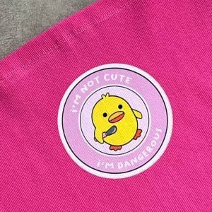 یک تاپ یا کراپ زنانه صورتی با بندهای نازک اسپاگتی. در مرکز سینه تاپ، یک پچ دایره‌ای با پس‌زمینه صورتی کم‌رنگ و حاشیه سفید دیده می‌شود. در مرکز این پچ، تصویر یک جوجه کارتونی زرد رنگ قرار دارد که یک چاقوی زرد در دست گرفته است. بالای جوجه، متن "I'M NOT CUTE" و پایین آن "I'M DANGEROUS!" (من کیوت نیستم، خطرناکم!) نوشته شده است. پارچه تاپ به نظر بافت‌دار و راه راه عمودی است و در پس‌زمینه یک سطح خاکستری دیده می‌شود از نمای نزدیک عکاسی شده.