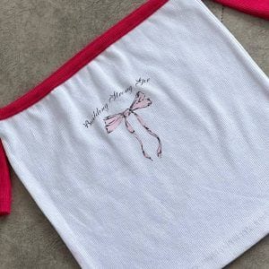 تاپ کراپ آف‌شولدر سفید و صورتی با طرح پاپیون صورتی و متن "Building Strong Girls" (ساختن دختران قوی)، مناسب برای استایل روزمره