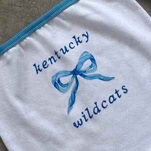 نمای نزدیک تاپ کراپ آف‌شولدر سفید و آبی با طرح پاپیون آبی و متن "Kentucky Wildcats"، مناسب برای طرفداران تیم یا استایل ورزشی