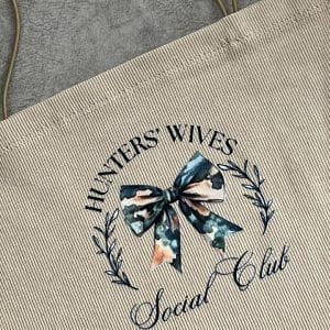 نمای نزدیک از تاپ کراپ زنانه با طرح 'HUNTERS' WIVES Social Club' و پاپیون گلدار در مرکز
